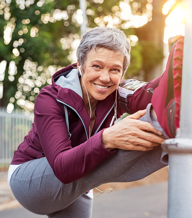 Smiling retired woman listening to music while stretching legs outdoors. Senior woman enjoying daily routine warming up before running. Sporty lady doing leg stretches and looking at camera.