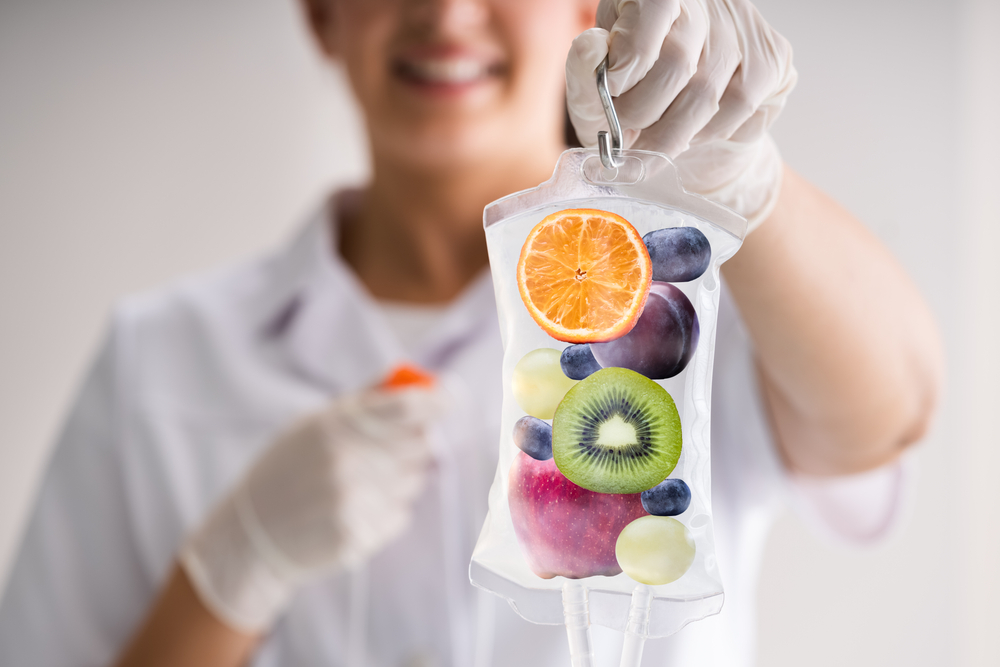 A naturopathic doctor smiles out of focus in the background while holding an IV bag filled with images of nutrient rich fruits and vegetables