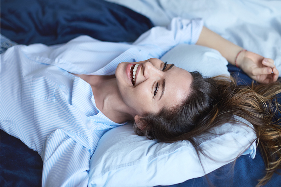 Attractive cheerful brunette female stretching in bedroom while lying in bed wearing blue shirt and jeans, laughing out loud with charming smile, full of energy, feeling excited about new happy day.